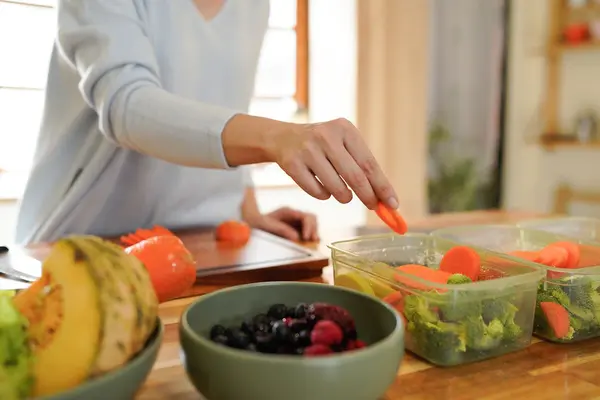 Eine Frau gibt Karottenstückchen in Meal Prep-Glasbehälter, in denen bereits anderes Gemüse liegt.