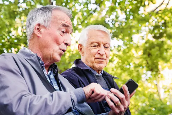 Zwei ältere Männer sitzen auf einer Bank im Park, einer zeigt dem anderen etwas auf seinem Smartphone.