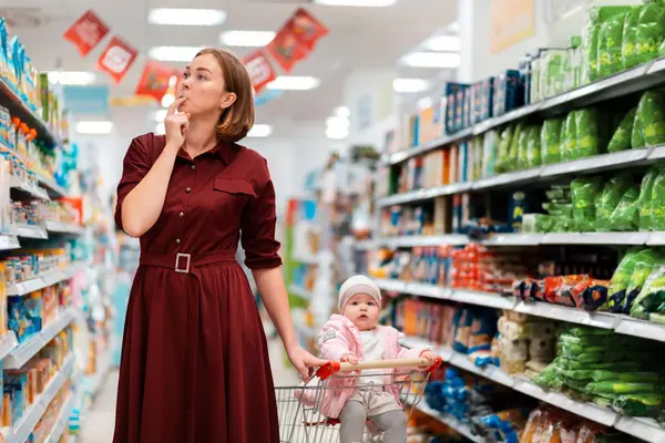 Eine Frau in dunkelrotem Kleid steht nachdenklich im Supermarktgang, den Finger an den Lippen, und blickt zu den Regalen hoch. Vor ihr schiebt sie einen Einkaufswagen, in dem ein Baby mit rosa Jacke und weißer Mütze sitzt."