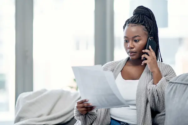 Dunkelhäutige Frau sitzt auf Couch mit Telefonrechnung in der Hand. Und telefoniert.