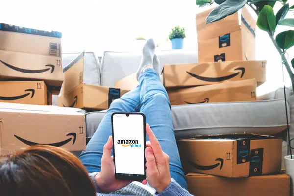 Eine Frau liegt auf dem Sofa mit dem Handy in der Hand, im Hintergrund ein Stapel mit Paketen von Amazon.