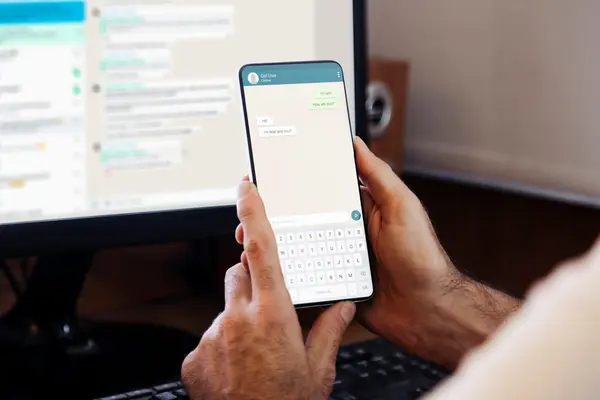 Smartphone in Hand, Messenger geöffnet, im Hintergrund angeschalteter Monitor