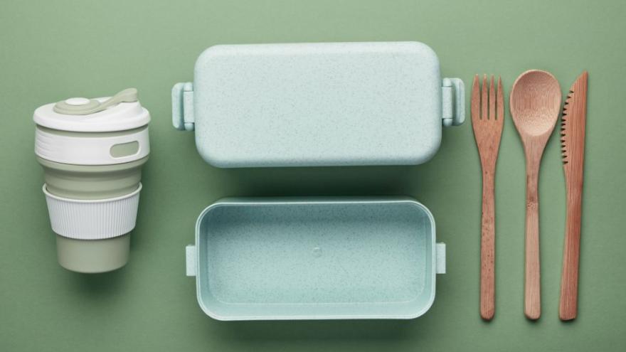 Foto von einem To-Go-Becher, einem Brotdose und Besteck aus Bioplastik und Holz, von oben fotografiert auf grünem Untergrund.