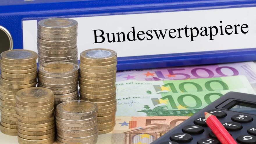Zu sehen sind aufgestapelte Euromünzen, ein Taschenrechner und ein Aktenordner mit der Aufschrift Bundeswertpapiere
