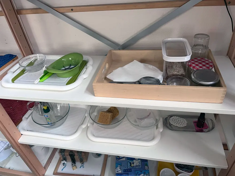 Regal in einer Kita, in der Tabletts mit den Materialien für kleine Alltags-Experimente stehen. Im Fokus stehen zwei Tabletts mit Experimenten rund um Ernährung: Eines mit einem Schneinebrettchen, einem Sparschäler, einem kleinen Küchenmesser und einem Apfelschneider sowie einer Glasschale. Das andere besteht aus Knäckebrot, mehreren Schraubgläsern, einem Teller, Servietten, einem feinmaschigen Sieb und Gläsern.