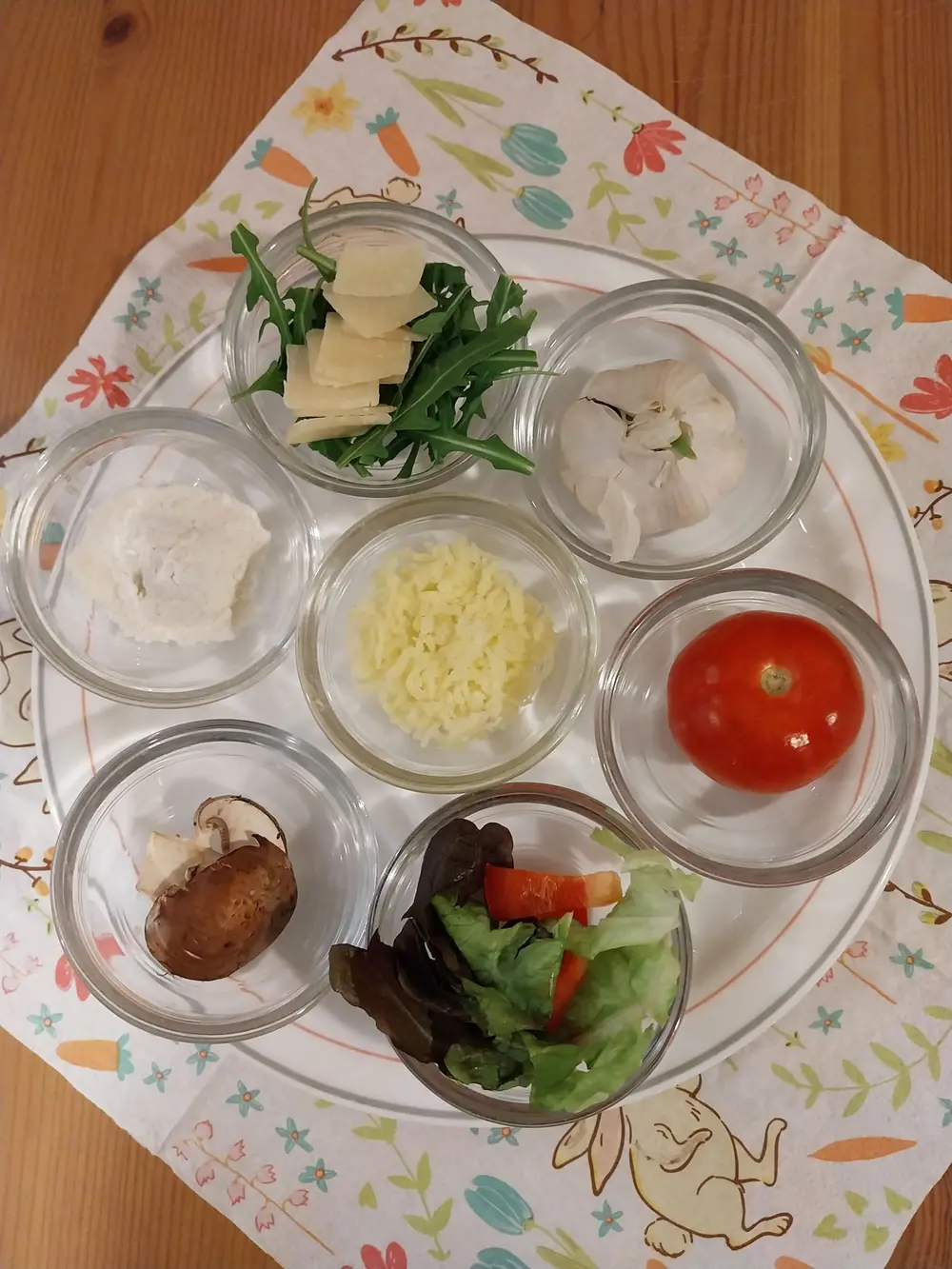 7 kleine Glasschälchen, die die Zutaten für eine Pizza (Mehl, Parmesan und Rucolasalat, Knoblauch, Reibekäse, Champignons, Tomate) und einen Beilagensalat (Blattsalatblätter und Paprikastreifen) enthalten.