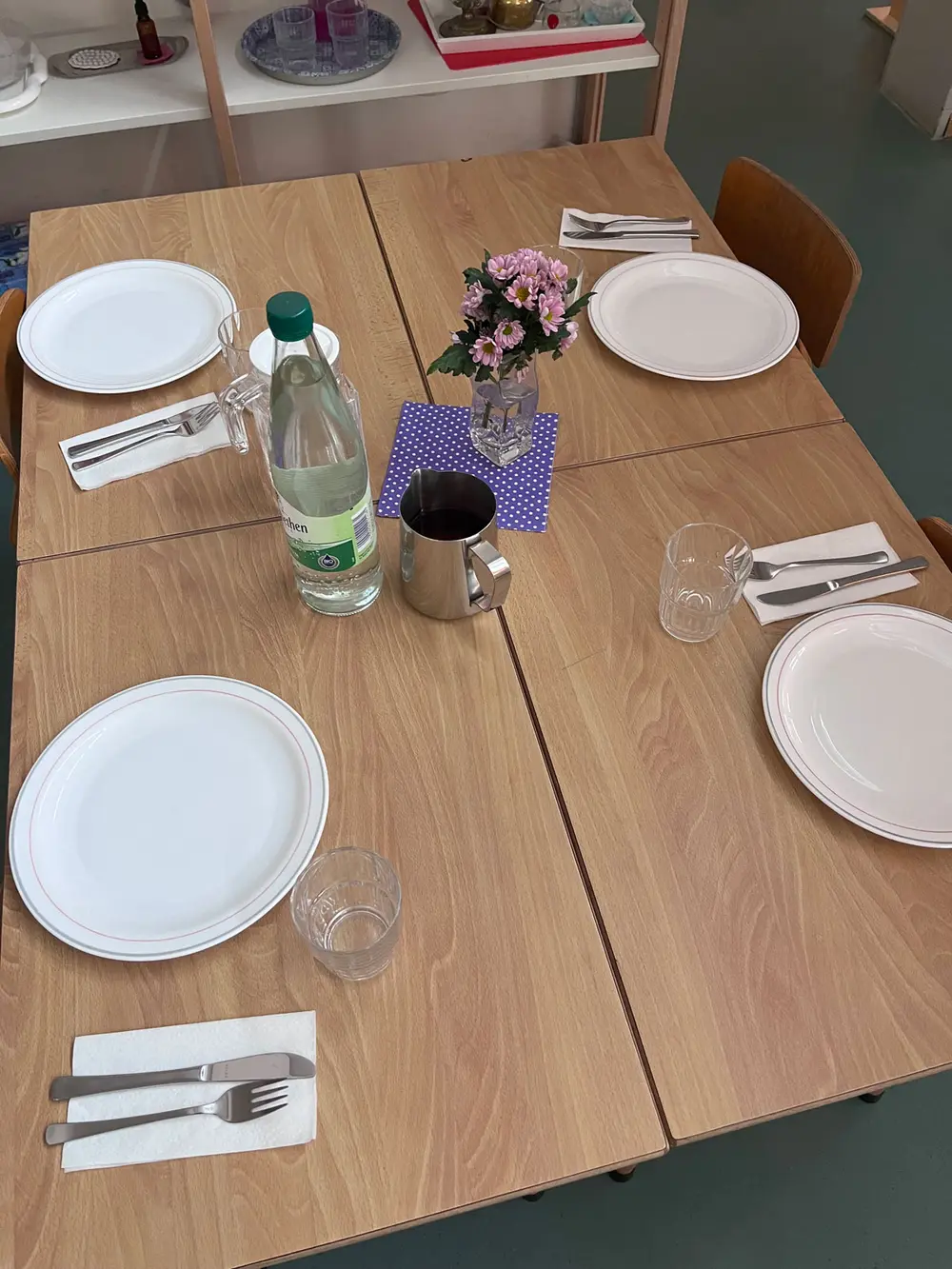 Gruppentisch für 4 Kita-Kinder mit Tellern, Besteck und Gläsern eingedeckt und einer Wasserflasche, einer Wasserkaraffe und Dekoration aus frischen Blumen und einer bunten Serviette.