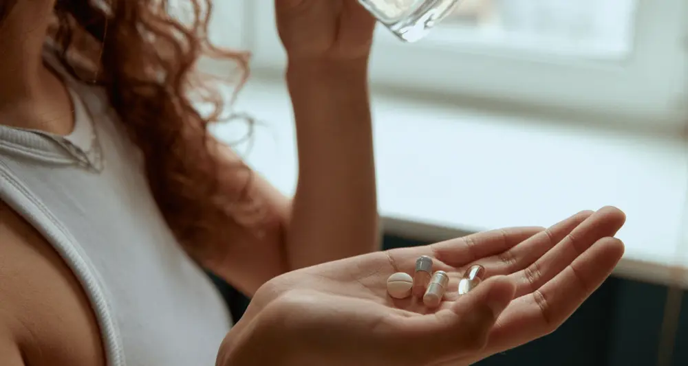 Person trinkt aus einem Glass und hält Tabletten und Kapseln in einer Hand