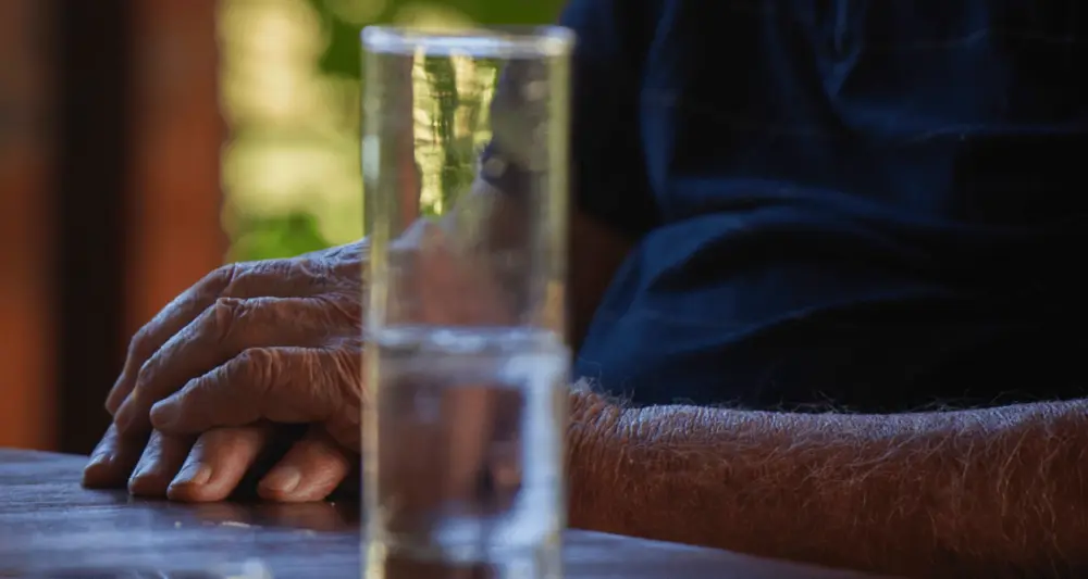 Zusammengefaltete Hände auf einem Tisch, unscharf im Vordergrund ein Glass Wasser