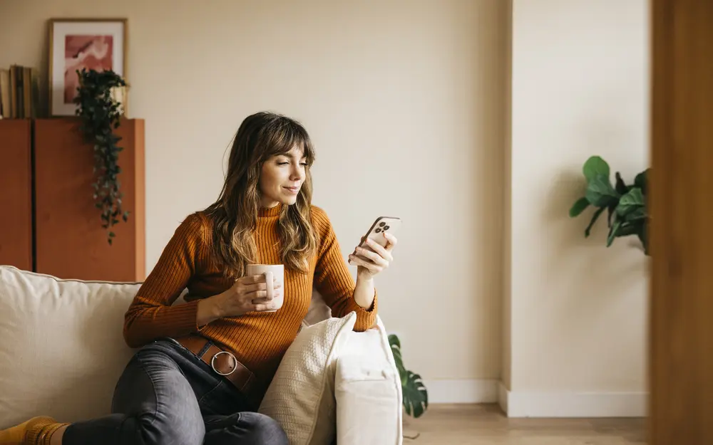 junge Frau auf dem Sofa mit Handy und Kaffeebecher in der Hand, zufrieden