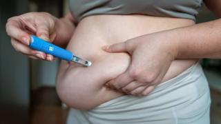 Eine Nahaufnahme zeigt eine Person, die sich mit einem blauen Injektionspen (ähnlich einem Insulin- oder Abnehmspritzen-Stift) ein Medikament in das Bauchfett injiziert. Die Person trägt Sportkleidung und drückt mit einer Hand eine Hautfalte am Bauch zusammen, während sie mit der anderen Hand den Pen senkrecht ansetzt. Der Hintergrund ist leicht unscharf und zeigt einen Innenraum.